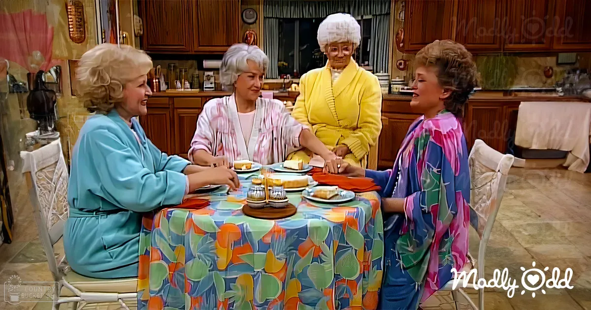 Four adult females in a domestic setting wearing brightly colored robes with simple necklines and no visible footwear; round table covered with a floral-patterned tablecloth, plates of food remnants, wooden cabinets, curtains, and kitchen items on the counter;