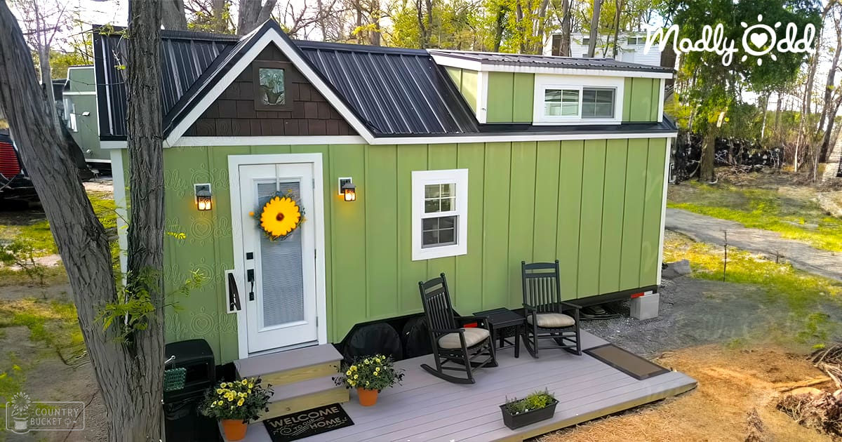A small green tiny house with a porch, surrounded by trees in an outdoor residential area.