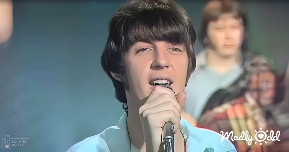 A young adult male singer wearing a light blue collared shirt performs on stage with a microphone, accompanied by another individual in a white patterned top.