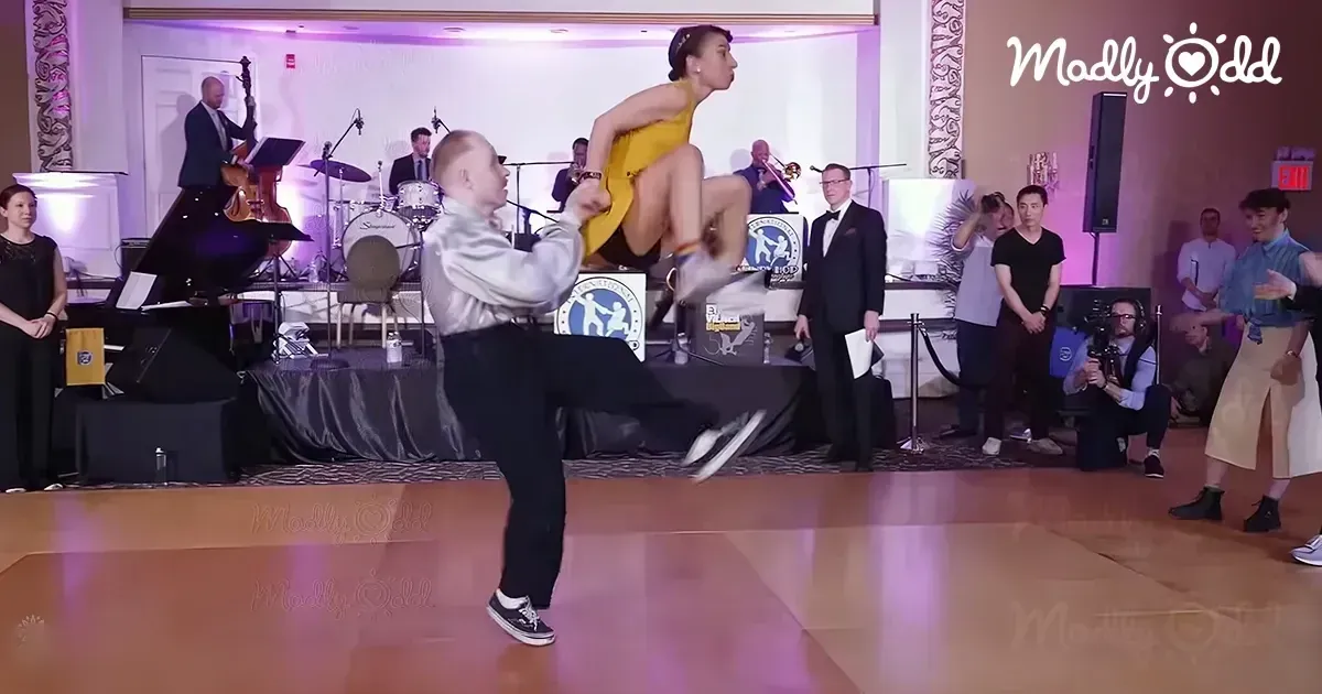A dance performance in a lively indoor event space with a male adult dancer in white and black attire and a young female dancer mid-air in a vibrant yellow dress, surrounded by an audience of various ages.