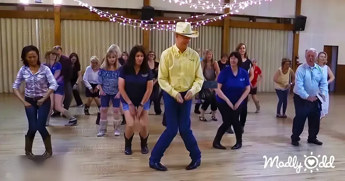 A group dance class in an indoor community hall with wooden floors, string lights, and a plain wall background.