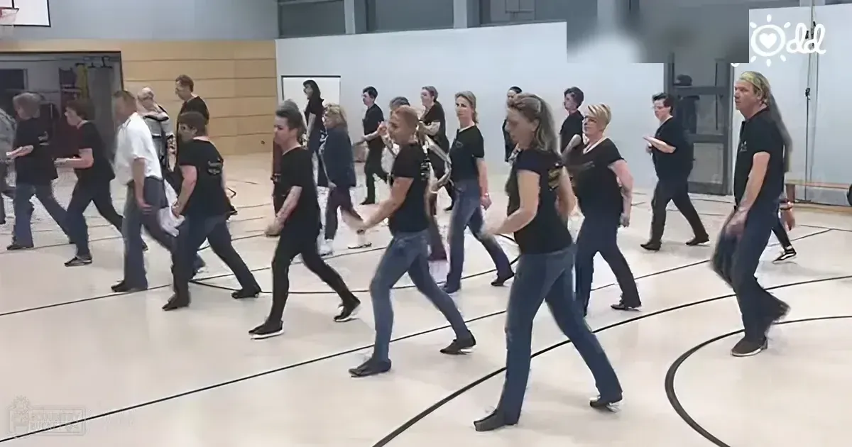 Group dance class in an indoor gymnasium with adults wearing black t-shirts and casual athletic shoes, set against a white wall and door backdrop under artificial lighting.