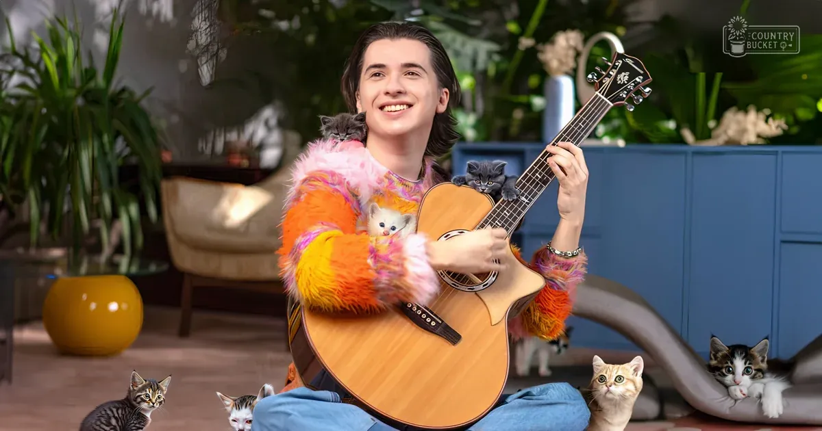 A young adult male playing guitar surrounded by tabby cats in an indoor setting with greenery, furniture, and soft lighting.