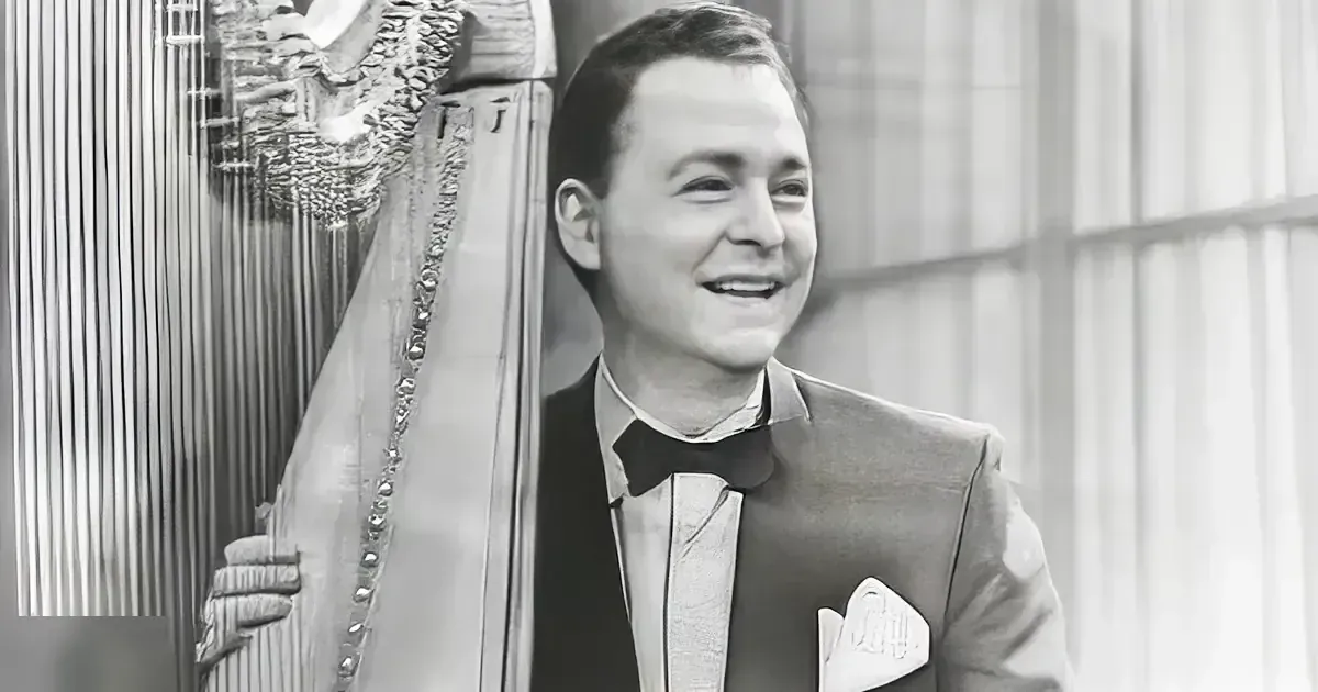 A young adult male stands indoors wearing a dark tuxedo with a bow tie, white dress shirt, and pocket square, likely at an upscale event.