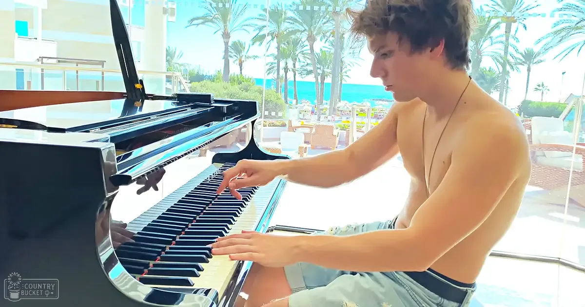 A young adult male plays a grand piano on an outdoor patio overlooking a beach with palm trees and modern building structures in the background.