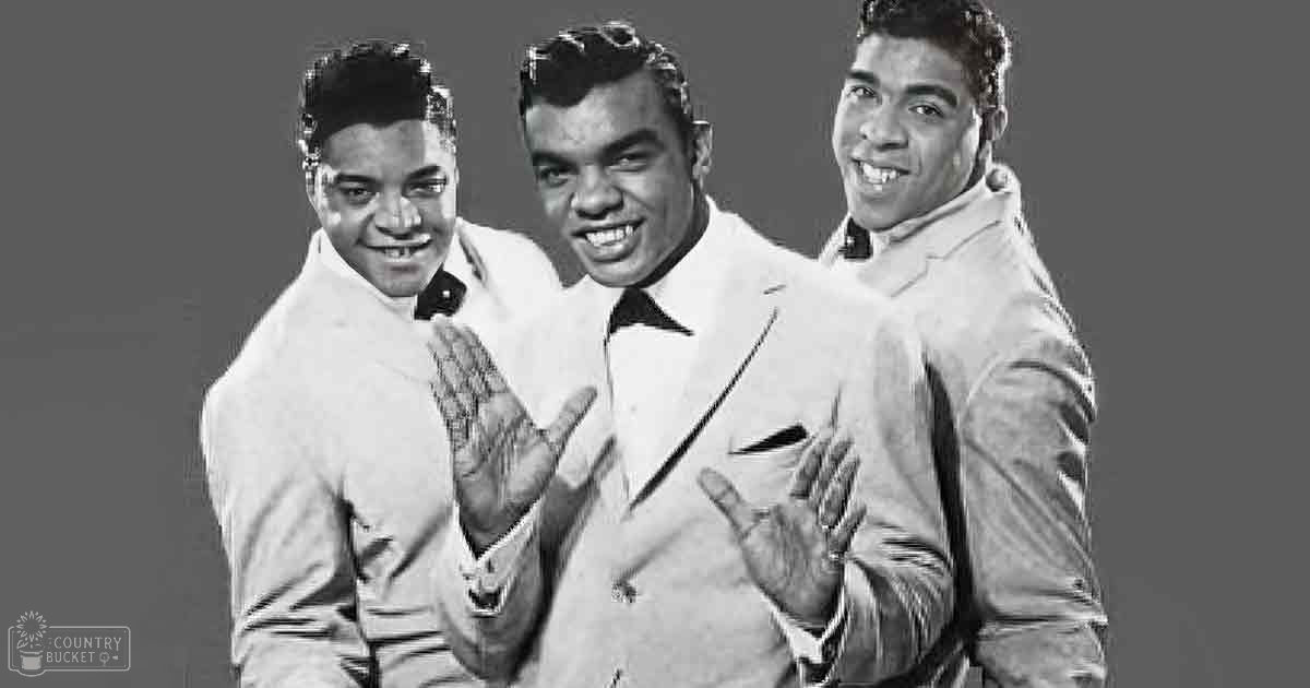 A mid-20th century studio group photo with three young adult males wearing formal suits and ties, set against a plain neutral background under bright even lighting.