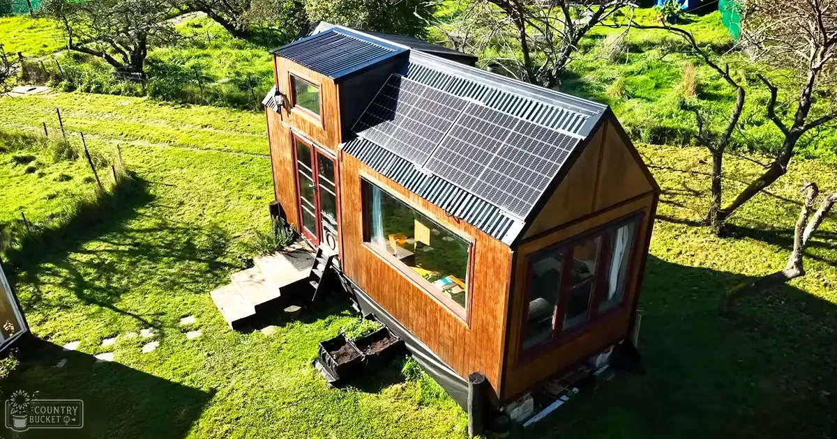 A small wooden house with large windows and solar panels, situated in a rural area surrounded by greenery on a sunny day during spring or summer.