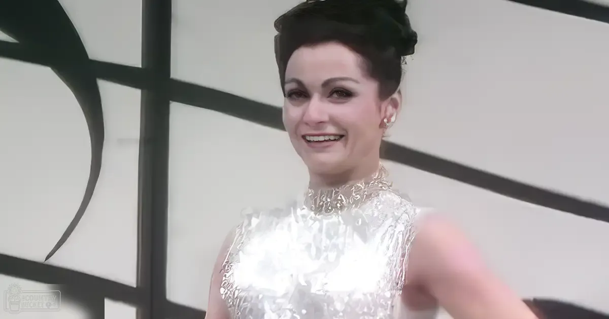 An adult female wearing a sleeveless, high-necked metallic silver dress with earrings, set against a modern indoor backdrop. Hand partially visible suggesting interaction.