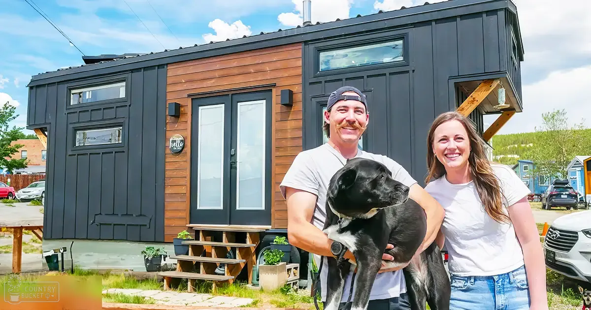 A Labrador Retriever dog with black coat and white markings stands in front of a small wooden house with a silver car parked nearby. The scene is outdoors, suggesting it might be spring or summer.