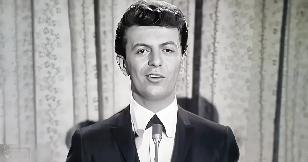 A young adult male wearing a dark suit jacket with a classic mid-20th century style stands in front of a microphone stand, set against a patterned curtain backdrop.