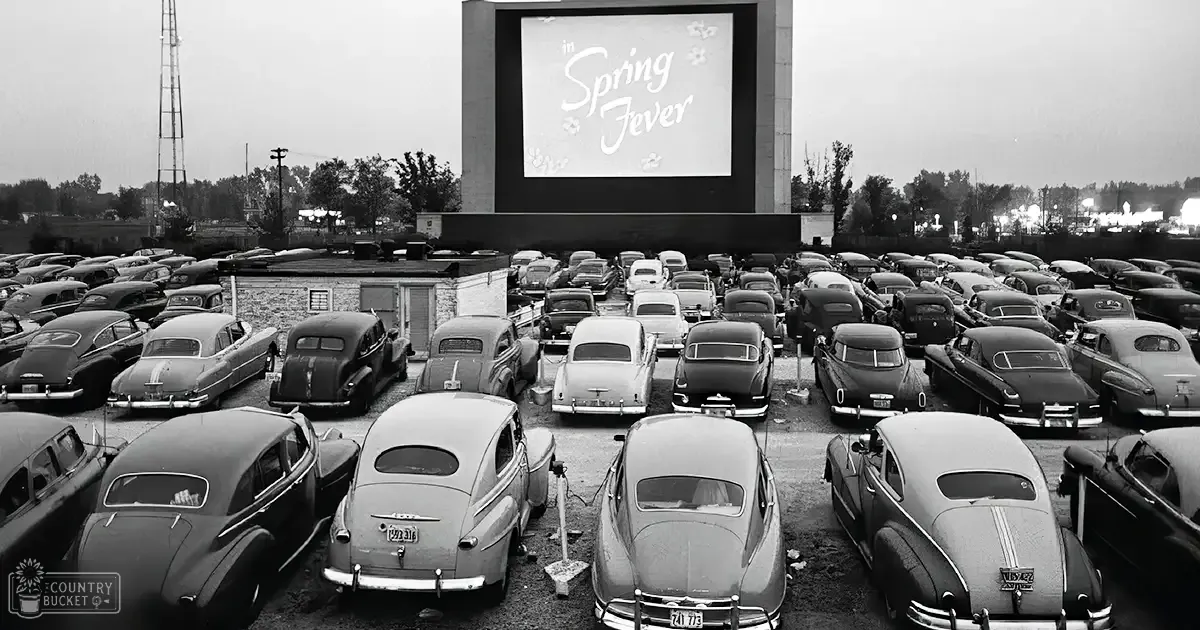 An outdoor drive-in movie theater with vintage cars from the 1950s era parked in rows facing a large screen displaying 'Spring Fever,' surrounded by trees and an industrial radio tower, likely during evening or early night time.