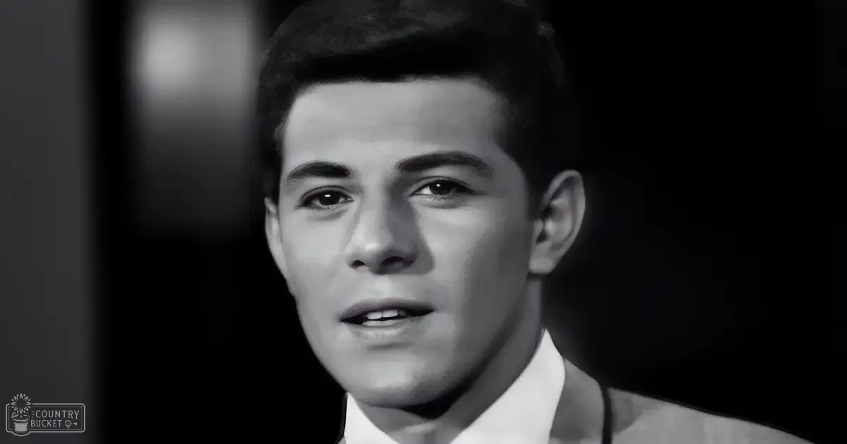 A young adult male wearing a white collared shirt with a dark jacket over it stands indoors in dim lighting, possibly evening or night.