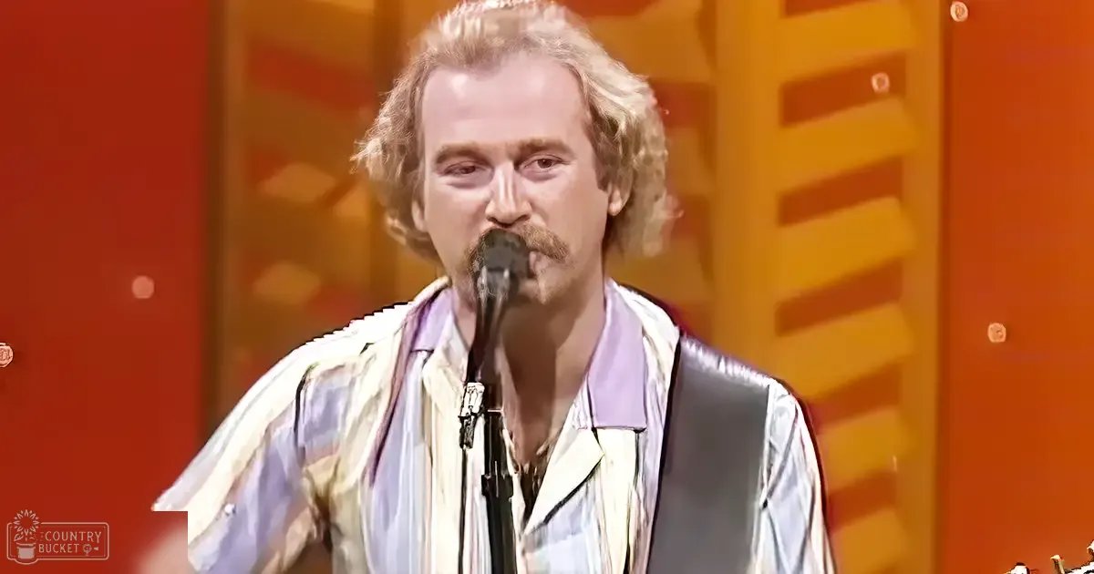 A middle-aged male performer wearing a patterned plaid shirt, sleeveless vest, and tie stands on stage with a microphone stand in the foreground under warm orange lighting.