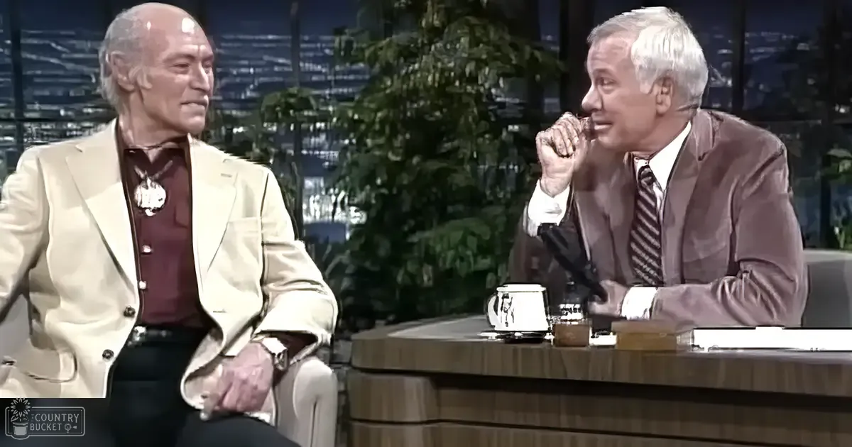 An indoor talk show setting with older adult males, wooden desk, mug, papers, potted plant, urban night scene through large windows, late fall or winter atmosphere.