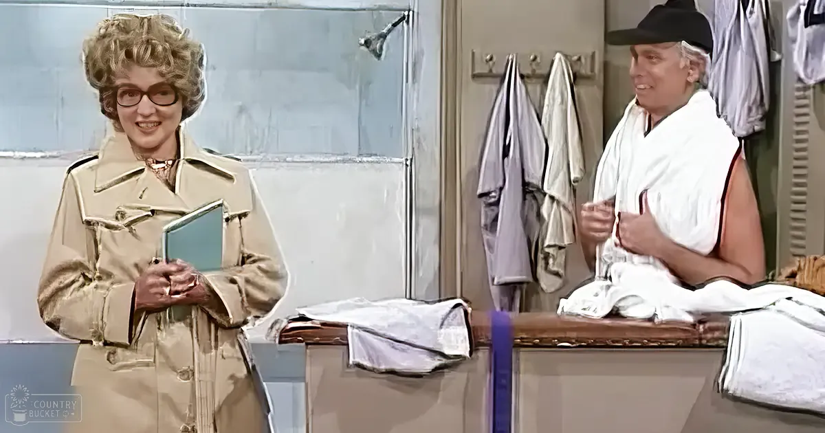 An older adult female wearing a beige trench coat stands indoors, possibly in a locker room with towels and shirts hanging around.