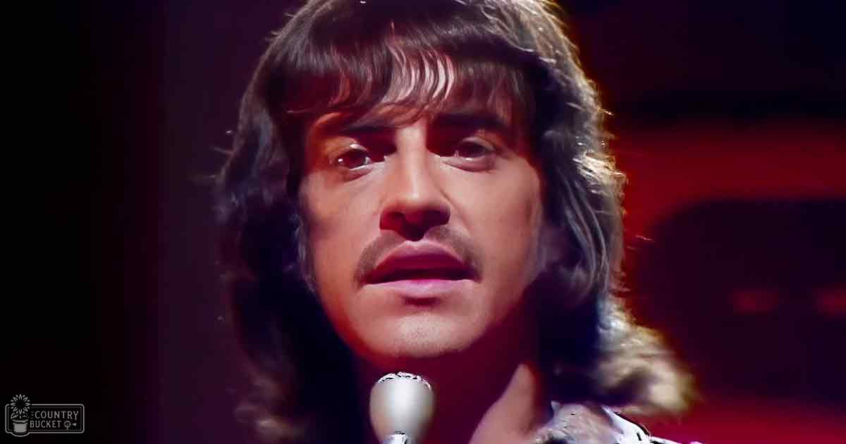 Adult male performer in relaxed, long-sleeved white shirt with floral or paisley pattern, set against red-lit stage background suggesting a performance venue.