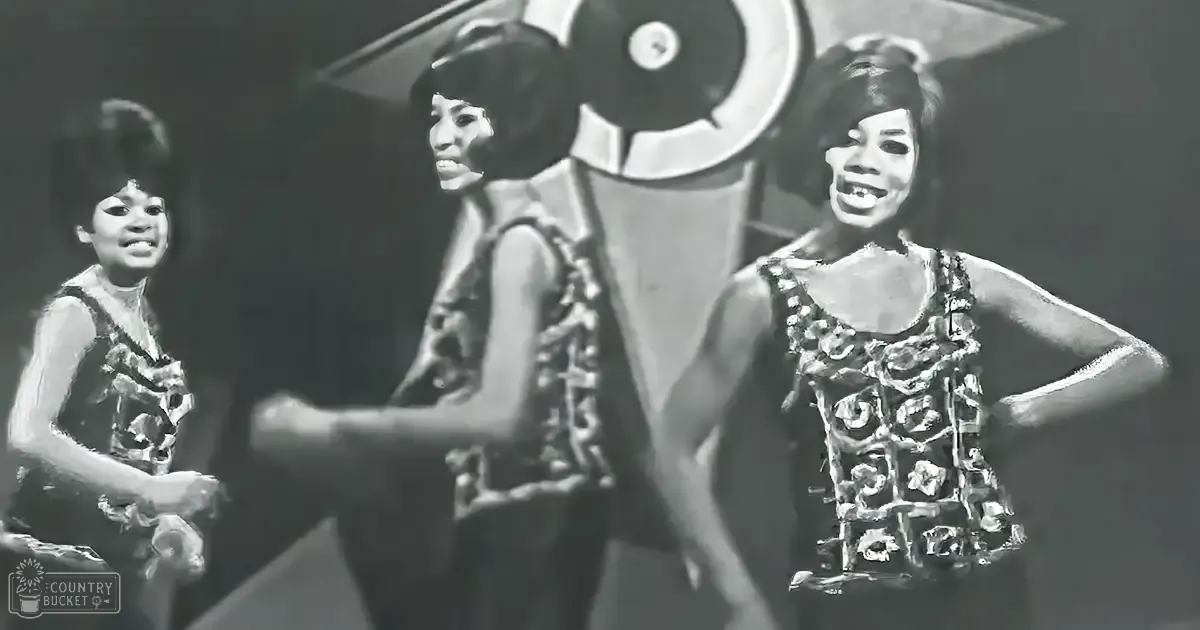 A musical performance scene from the 1960s with three young female performers wearing floral-patterned dresses on a dimly lit stage.