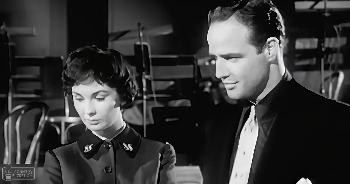 A mid-20th-century indoor setting with a young adult female wearing a dark jacket with decorative buttons and a middle-aged male in a formal suit, possibly from a film or television show.