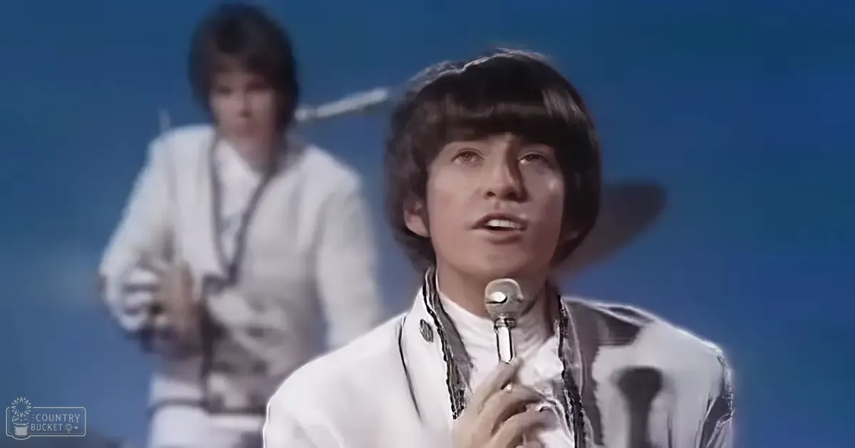 A young adult male singer in a white jacket with black trim, performing on an indoor stage with blue lighting. Another individual is visible in the background holding a microphone.