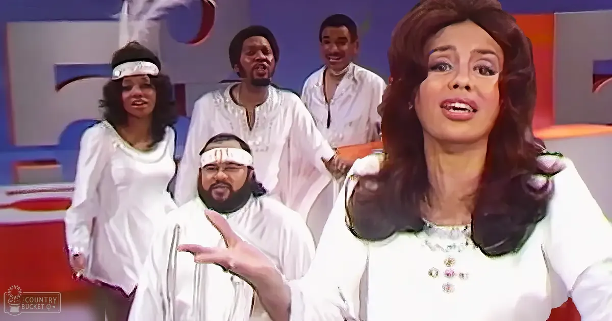 Adult female with long brown hair styled in waves, wearing a white garment adorned with rhinestones or jewels at an indoor event or performance.