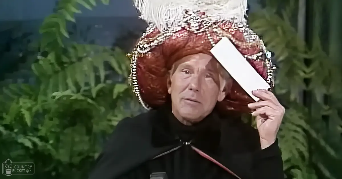 An older adult male wearing a large, ornate red and gold-adorned hat with feathers and a black woolen robe stands indoors amidst lush greenery. The setting suggests a theatrical or performance environment.