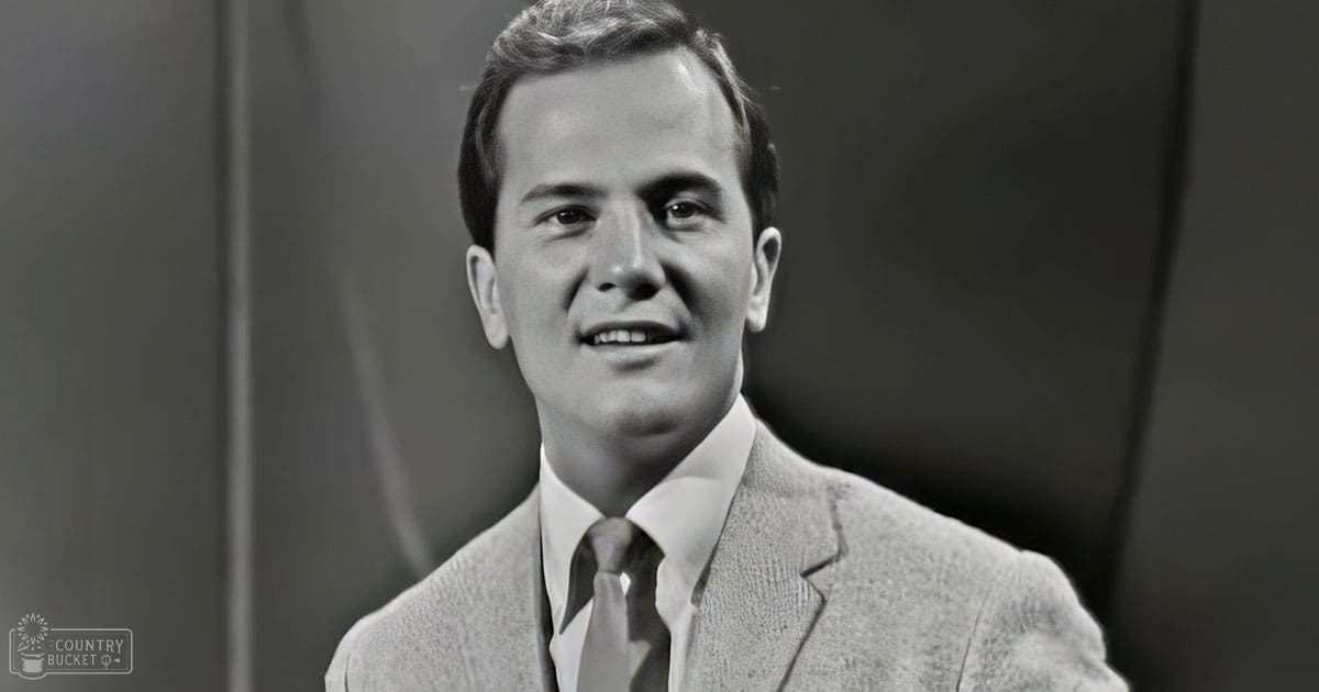 A young adult male in a tailored suit jacket with textured fabric, possibly wool or tweed, stands indoors against neutral-colored walls with subtle curves. The setting is simple and professional, with even lighting suggesting daytime.