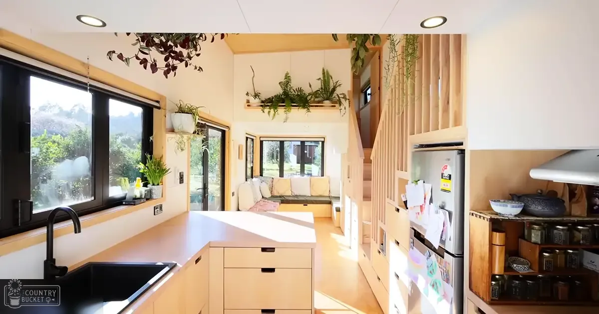 A cozy interior of a modern tiny house with light wood cabinets, dark-framed windows, and hanging plants in a temperate region during daytime.