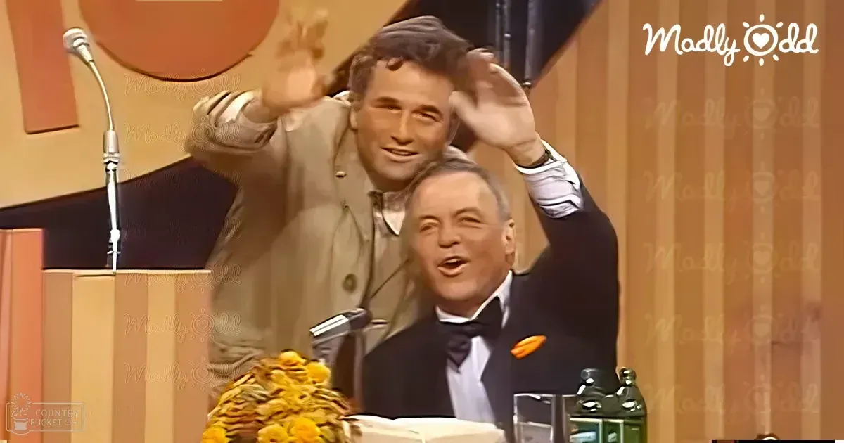 A television show set in an indoor studio environment with two middle-aged men, one standing and dressed casually in a light-colored jacket, and another seated wearing a dark suit with a bow tie. The setting includes a podium, microphone, yellow flowers, and a green bottle on the table.