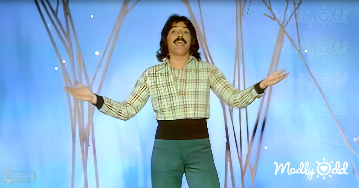 Adult male singer in a relaxed, casual fit long-sleeved light green plaid shirt and pants against a studio backdrop with blue gradient and abstract white lines and dots. Indoor setting under artificial lighting.