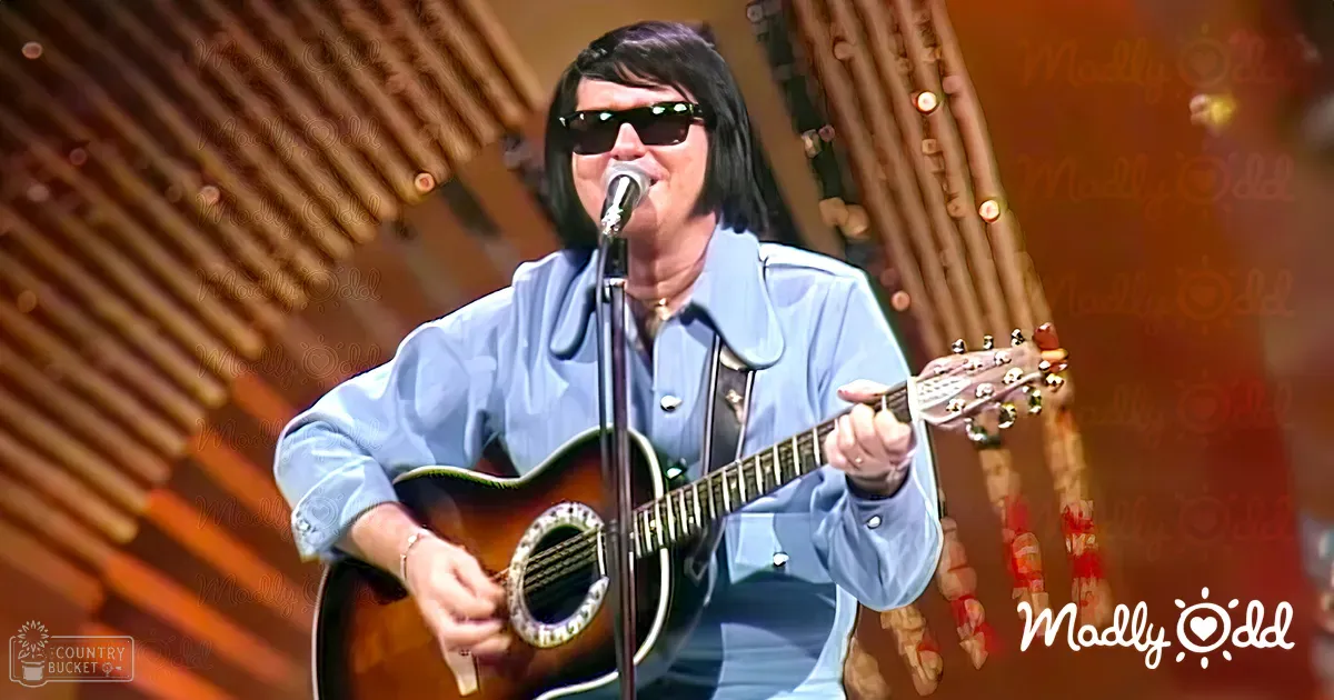 A male adult singer wearing a light blue, long-sleeved shirt with rolled-up sleeves, sunglasses, and a bracelet, playing an acoustic guitar on a stage during what appears to be a performance.