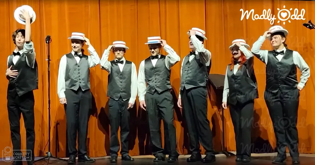 A group performance scene with young adult males in white shirts, black vests, bow ties, dark trousers, and white hats with red stripes, set against an orange curtain backdrop at a school auditorium during evening indoor lighting.