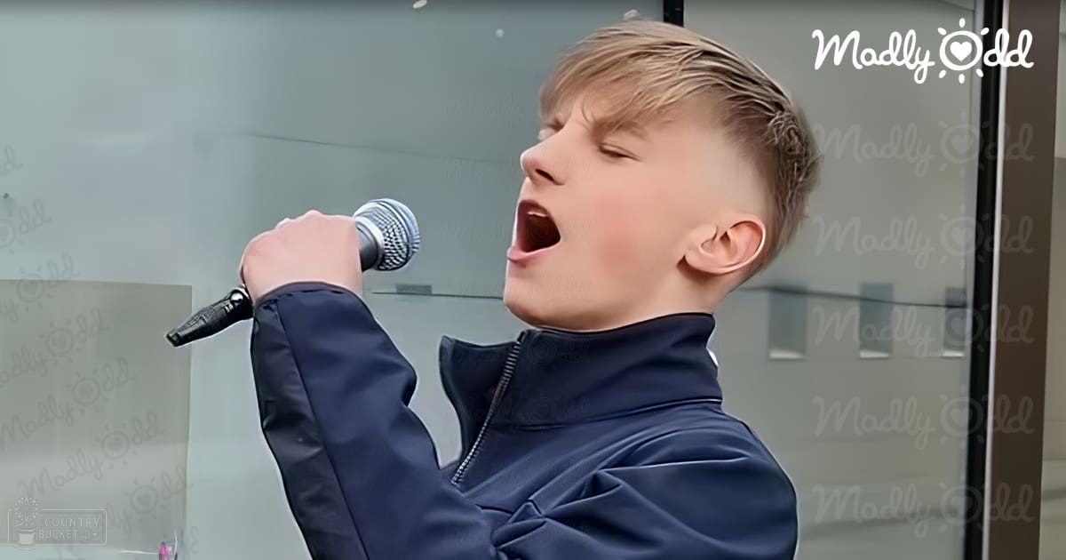 Young adult male singing into a microphone, wearing a dark blue zip-up jacket with high collar and relaxed fit indoors.