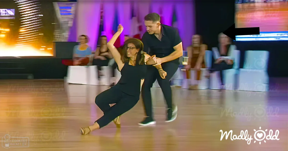 A dance performance in an indoor setting with white chairs and tables, purple lighting, and colorful visuals displayed on screens.