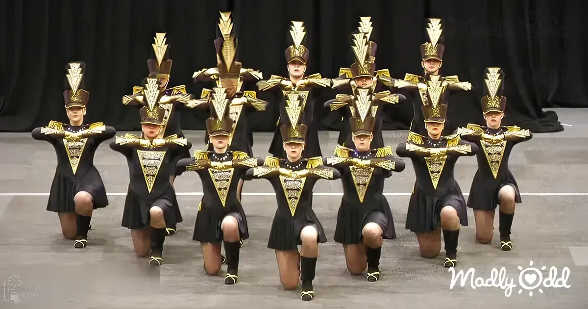 A group performance by young adult female dancers in a dance or marching band setting, wearing form-fitting uniforms with black and gold accents on stage under artificial lighting.