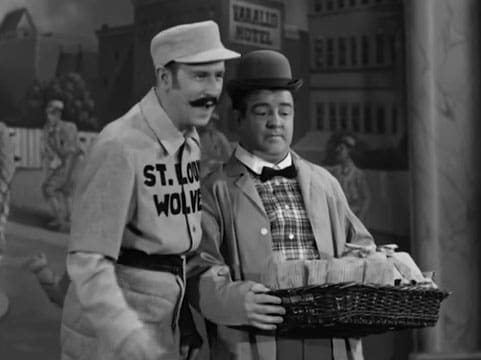 Two adults in a St. Louis Wolves baseball uniform interact on an urban street scene with buildings and a 'VARALIO MOTEL' sign visible.