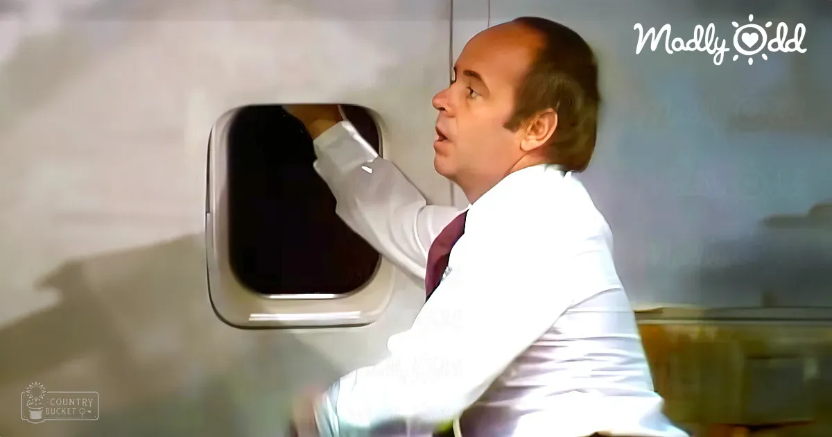 A middle-aged man wearing a white dress shirt with a maroon tie sits inside an airplane cabin on a sunny day.