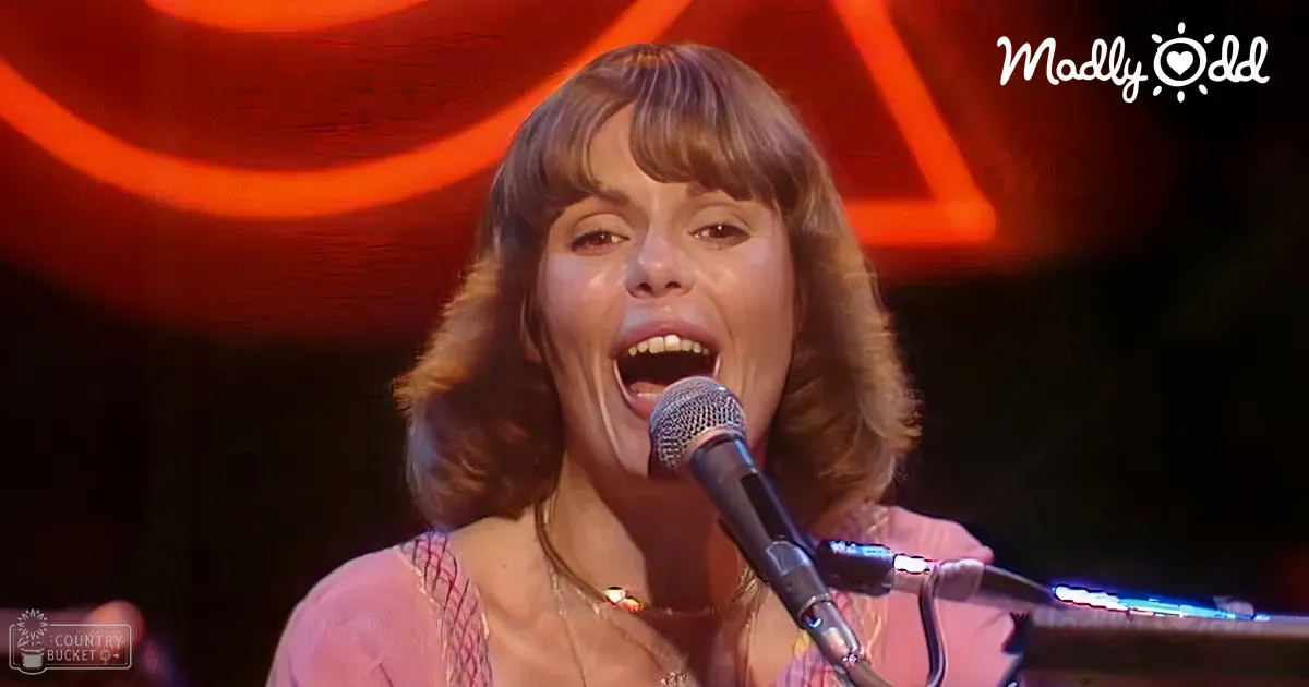 A middle-aged adult female singer performing into a microphone at an indoor concert venue with stage lighting.