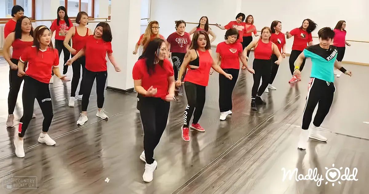A group fitness class in a well-lit indoor dance studio with adults wearing red t-shirts and black pants or leggings during daytime.