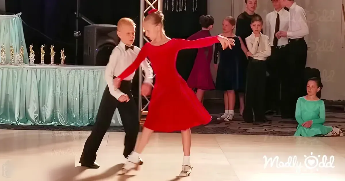 A young couple in formal dance attire dancing, likely at an event or competition. The setting appears to be indoors with spectators dressed formally.