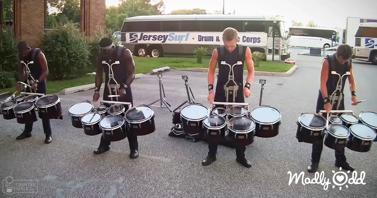 Adult males in sleeveless vests practicing a drum line at a parking lot with buses labeled "Jersey Surf Drum & Bugle Corps."