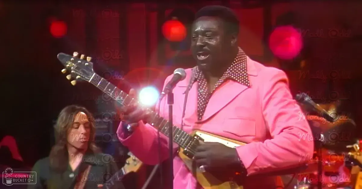Adult male performer in pink sequined suit jacket singing into microphone during stage performance with dim red lighting and text overlay 'Madly Odd'.