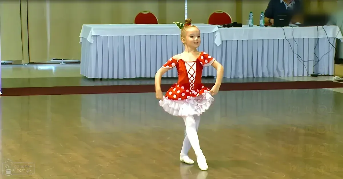 A toddler in a red and white polka dot ballet costume performing on stage with ruffled skirt, round neckline, short sleeves, and white ballet slippers. Decorative leaf accessory adorns her hair bun.