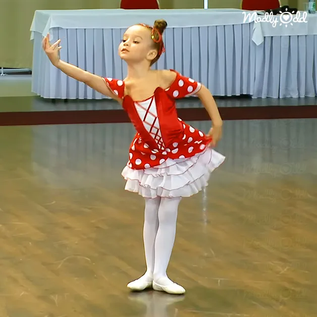 A young female child in a red and white polka dot leotard, white tutu skirt, tights, ballet shoes, and headband, dancing in an indoor dance studio with wooden or laminate flooring under bright fluorescent lights.