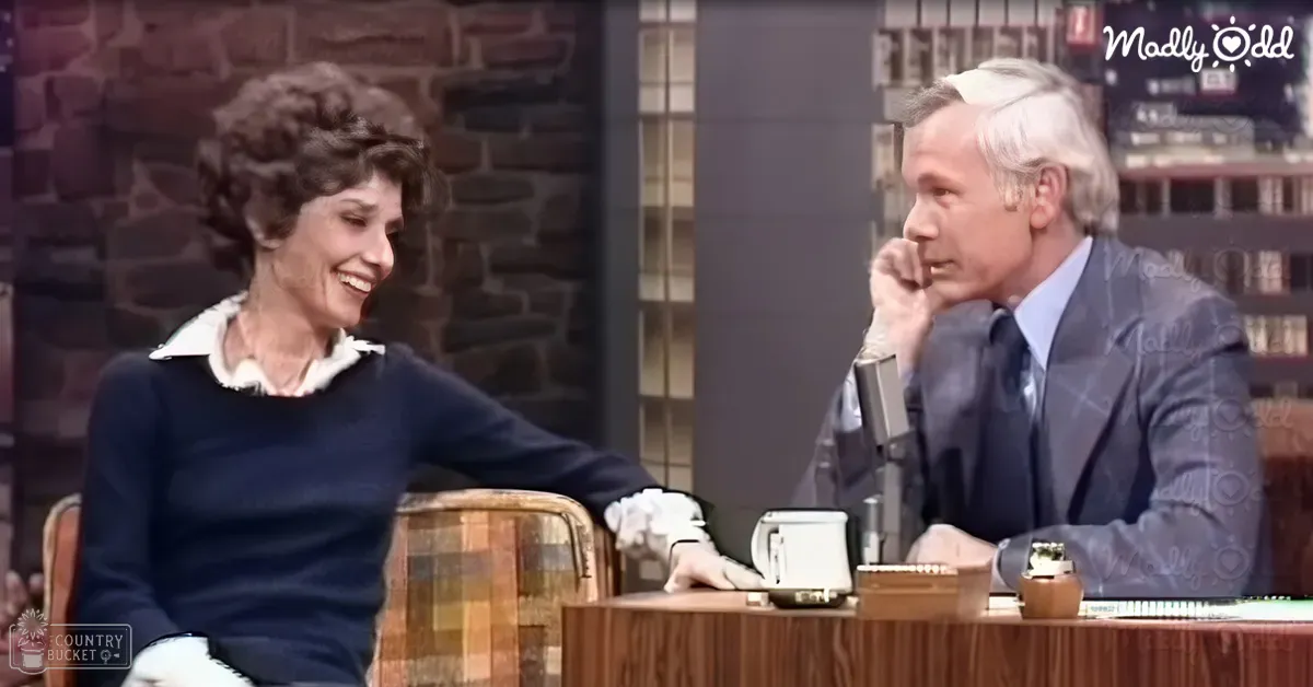 An older adult female wearing a dark blue vintage sweater with white accents sits on a wooden chair, engaged in an interview or talk show segment set against a rustic brick wall backdrop.