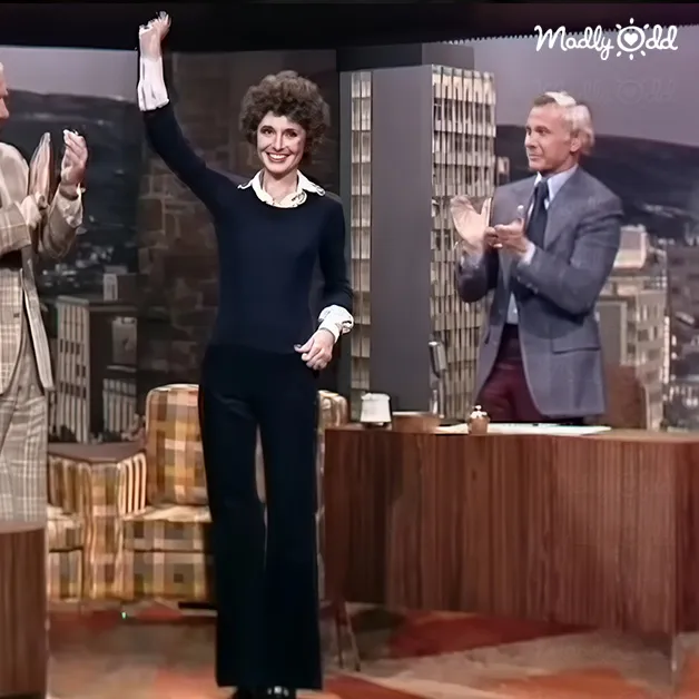A woman in her adult years wearing a black pantsuit with white accents and gloves, seated on a talk show set with other adults dressed formally. The backdrop suggests an urban high-rise studio design.