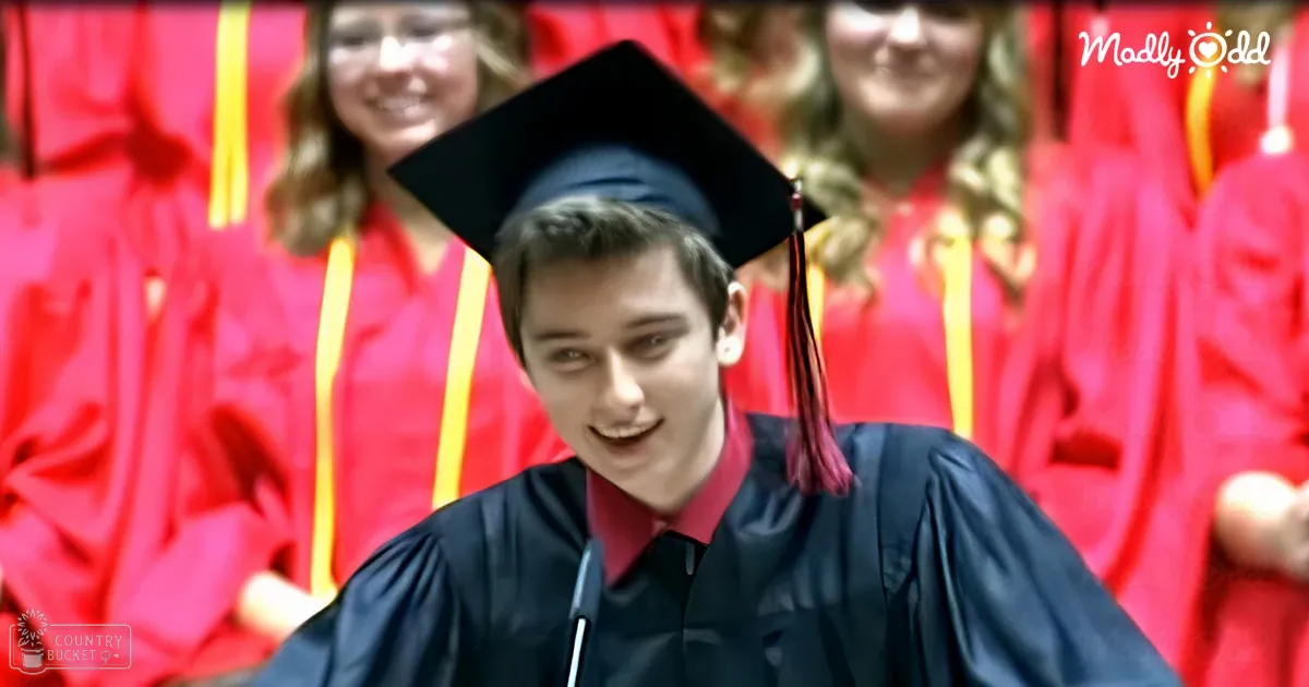Young adult male in a dark blue graduation gown with red tassel, seated among other graduates at an indoor school auditorium during a formal ceremony.
