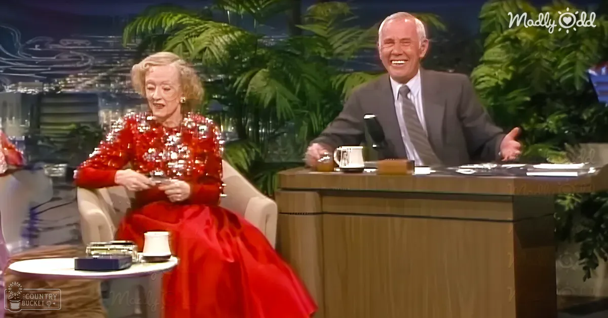 A middle-aged male talk show host in a dark suit and striped tie sits next to an older adult female guest wearing a vibrant red sequined dress, both seated at a wooden desk in a television studio with lush green plants and a 'Madly Odd' logo visible.