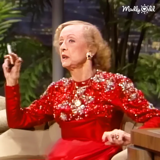 A middle-aged adult female in a form-fitting red sequined dress with full-length sleeves and large hoop earrings sits indoors on a light-colored couch surrounded by potted plants, with a wooden table holding what appears to be a glass of water nearby.