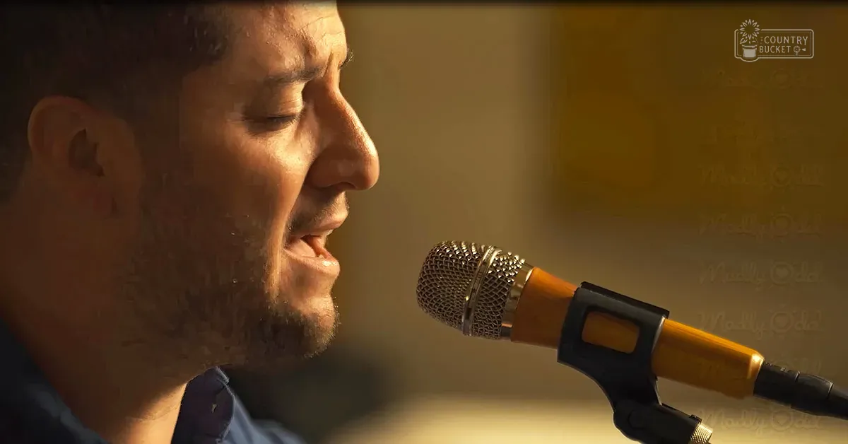 A middle-aged man wearing a blue shirt with long sleeves sings into an orange-microphone in a softly lit indoor setting.