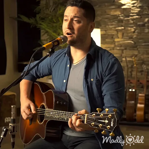 An adult male singer in a relaxed, casual long-sleeved button-up shirt over a t-shirt with a necklace, singing into a microphone while playing an acoustic guitar in a rustic indoor music studio or performance space.
